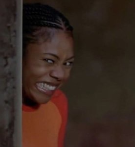 Woman with braids peeking around a wall with the most delightfully scheming grin