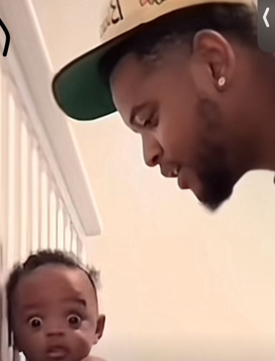 Wide-eyed shocked baby staring straight ahead as a man in a cap looks down at it