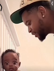 Wide-eyed shocked baby staring straight ahead as a man in a cap looks down at it