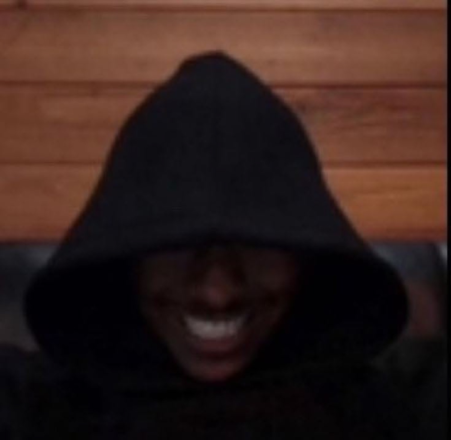 Person in a black hood shrouded in shadow with only a wide grinning smile visible