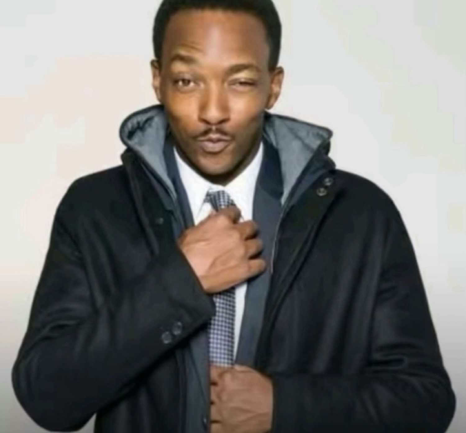 Man in a suit and dark coat gripping his tie with a smooth, flirty smirk