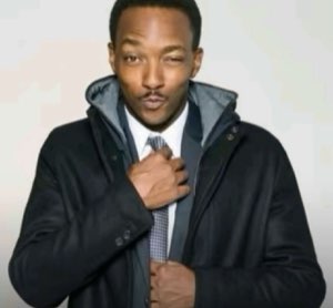 Man in a suit and dark coat gripping his tie with a smooth, flirty smirk