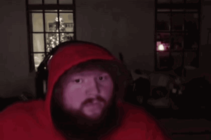 Bearded man in a red hoodie by a Christmas tree hiding his face in embarrassment