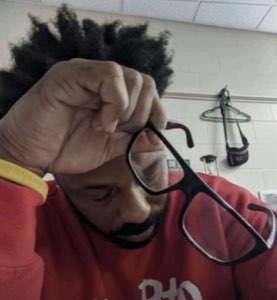 Man in a red shirt lowering his glasses and rubbing his eyes, completely worn out