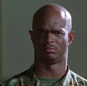 Bald man in camo fatigues staring down the camera with a tight, unimpressed frown