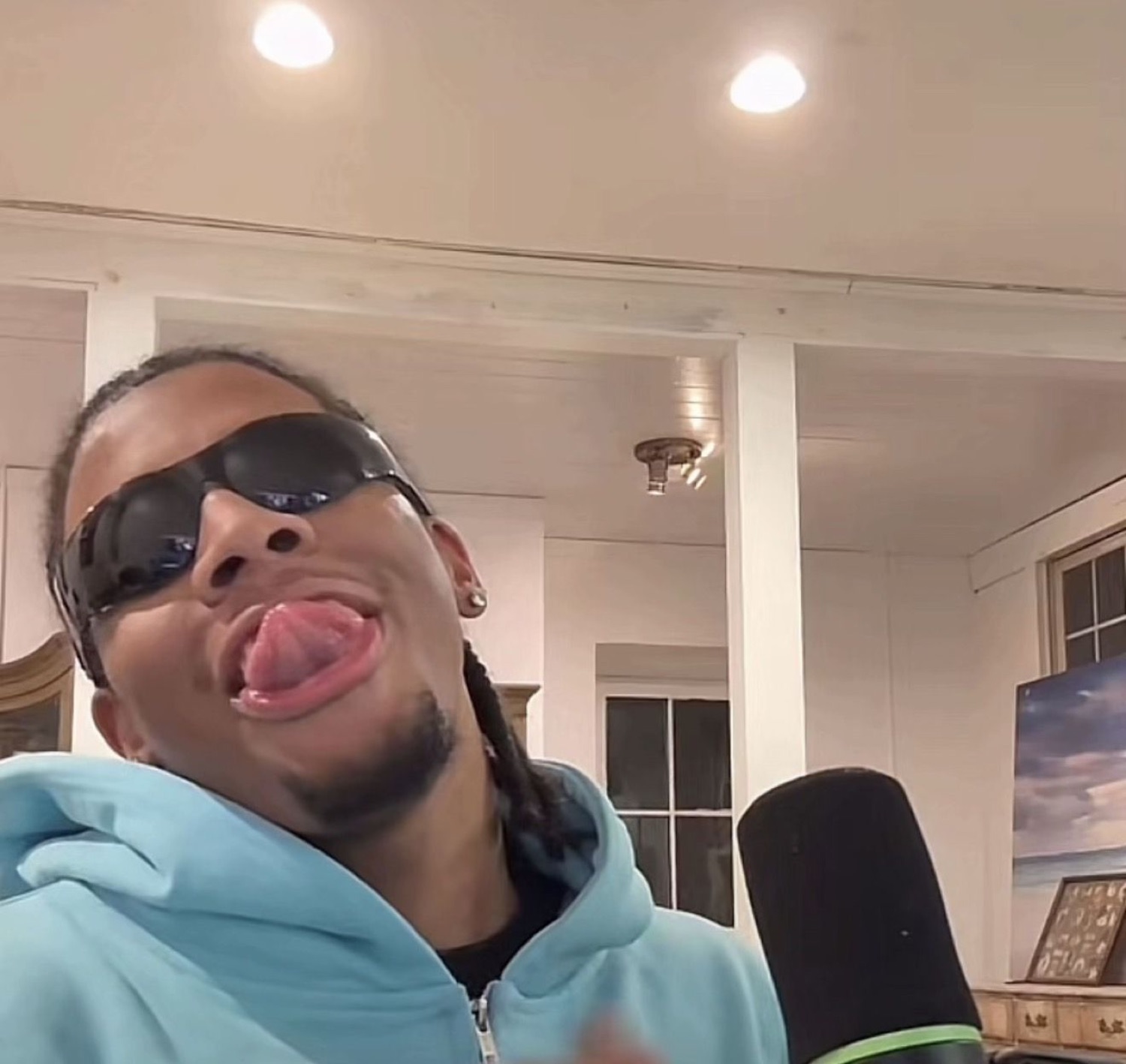 Guy in sunglasses and a hoodie sticking his tongue out into a mic, being goofy