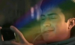 A man smiling softly with eyes closed at his phone, bathed in a rainbow glare