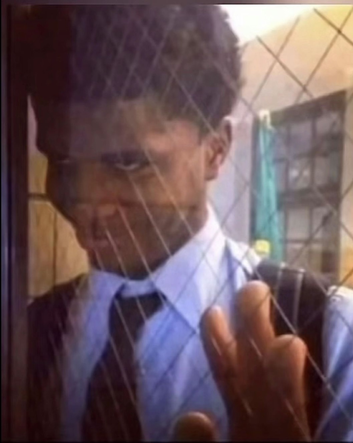 Boy in school uniform behind a chain-link fence giving a wary side glance