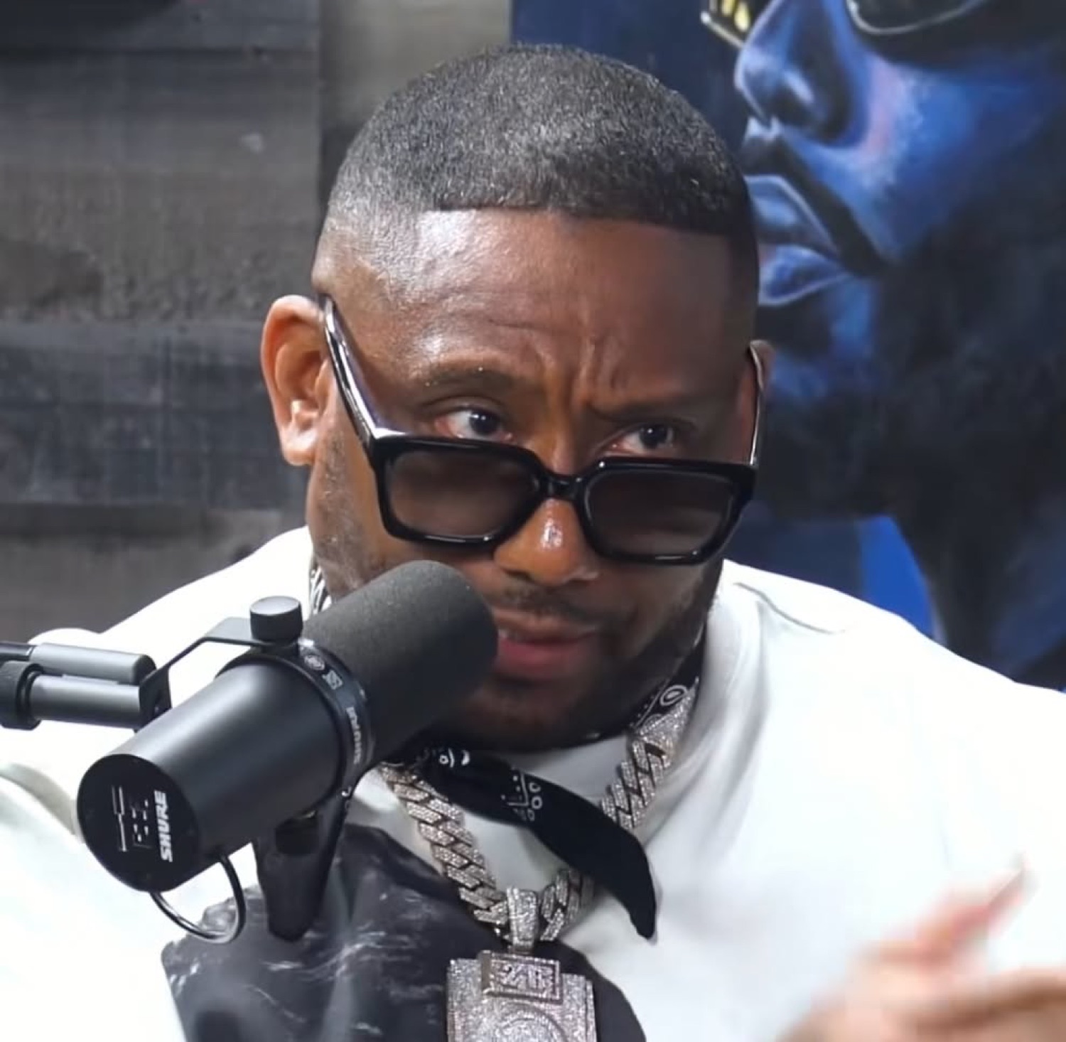 Man in black shades and a chain giving the podcast mic a skeptical raised brow