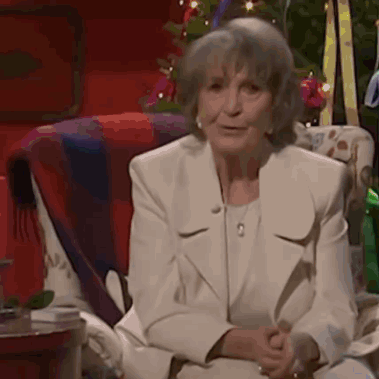 Elderly woman in a white suit by a Christmas tree doing the six seven hand gesture