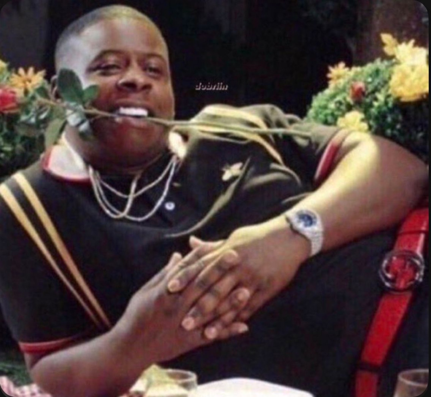 Man lounging confidently with a rose in his mouth, dripping in jewelry and a luxury watch
