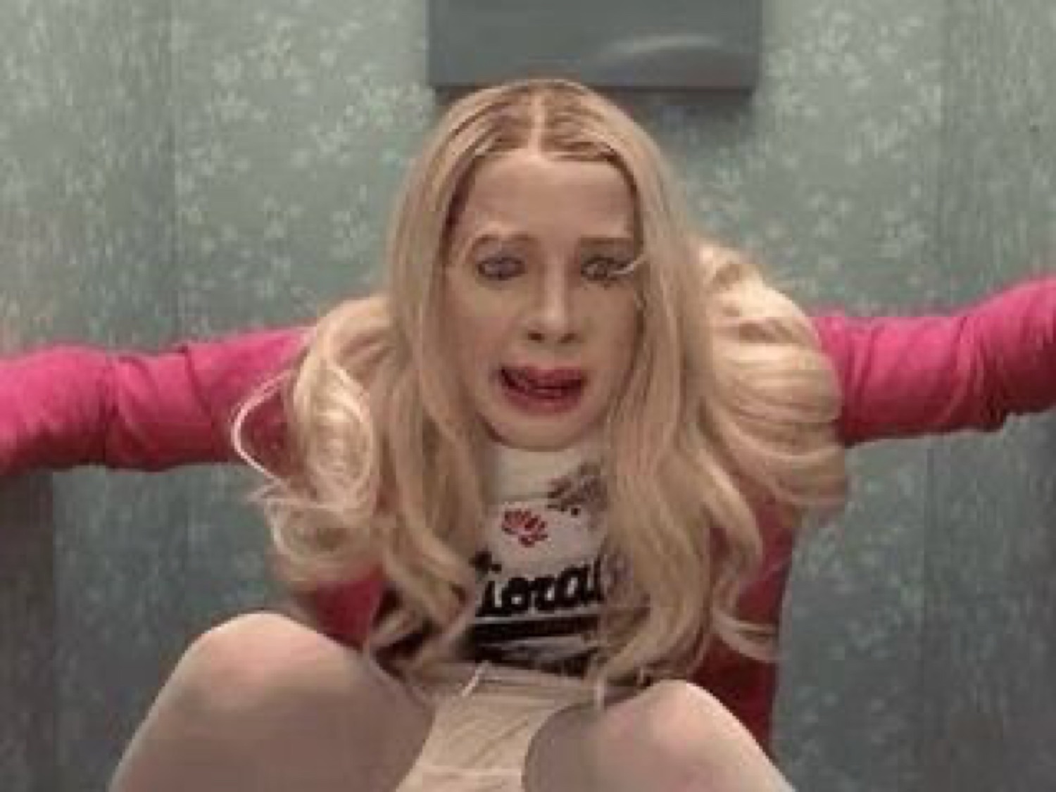 Person in a blonde wig and pink outfit sitting against a wall with a panicked, desperate expression