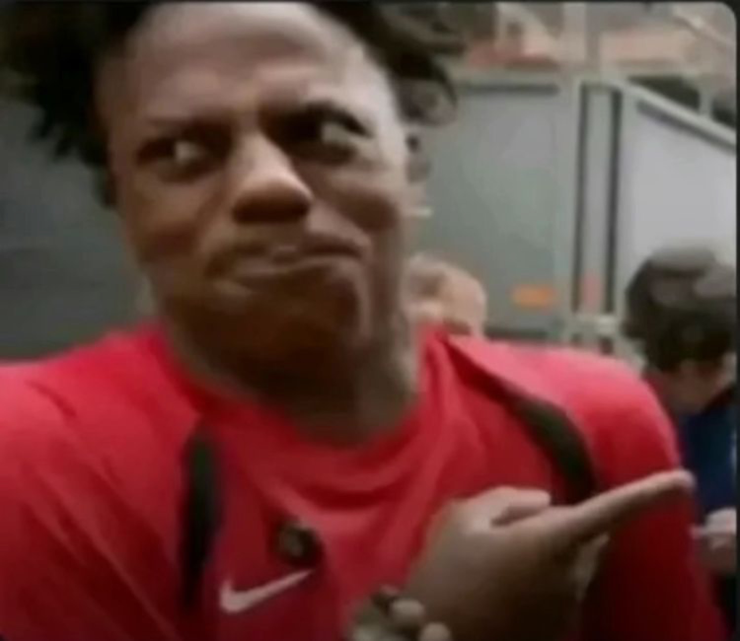Man in a red shirt side-eyeing and pointing off to the side with a skeptical face