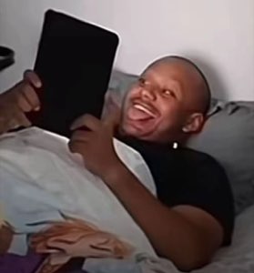 Guy lying in bed holding a tablet, mouth wide open laughing hysterically at the screen