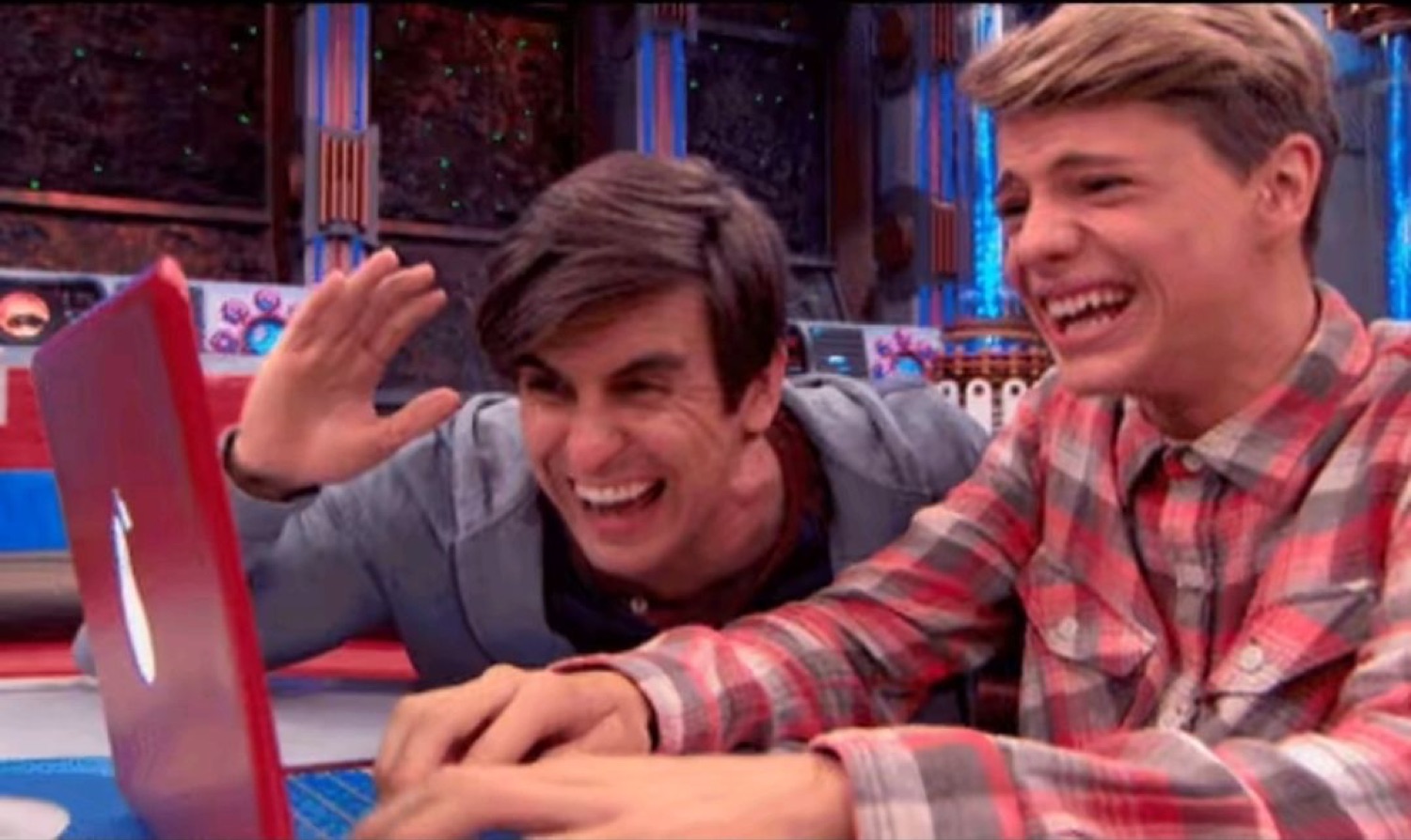 Two guys cracking up at a laptop, one waving his hand mid-laugh