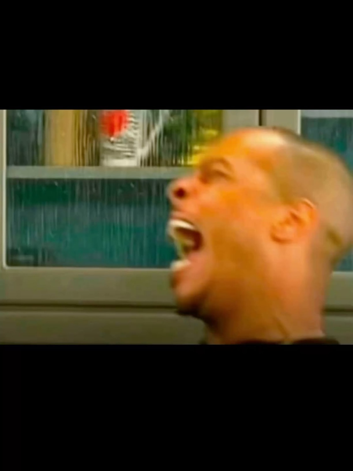 Man with a buzzcut screaming wide-mouthed in profile near a window