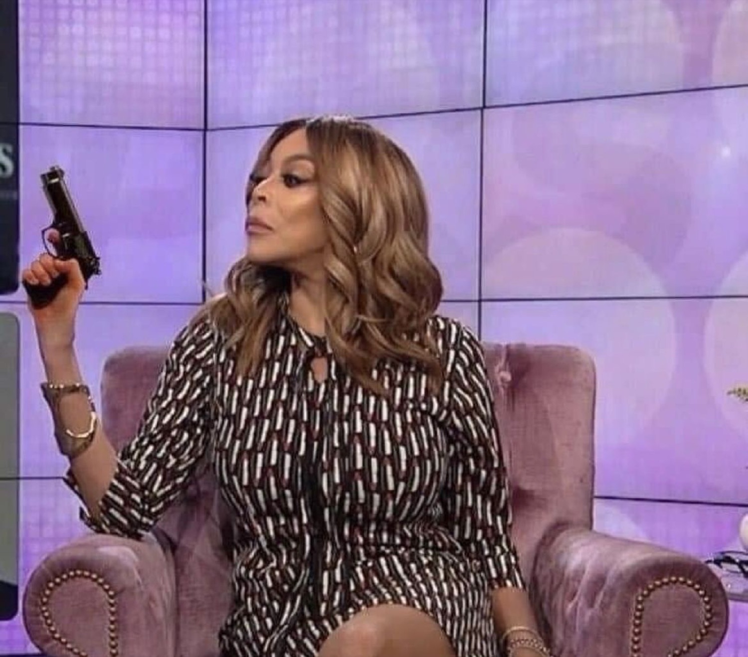 Talk-show host lounging in a chair casually holding up a handgun with a bored side glance