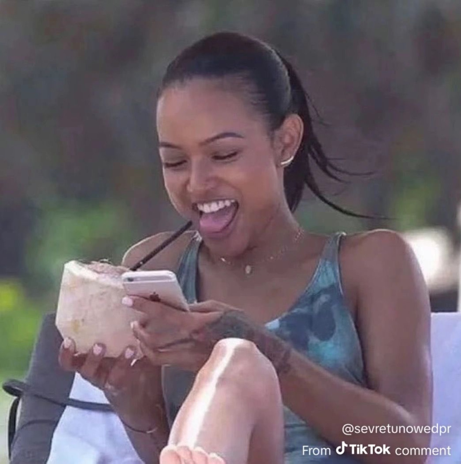 Woman laughing with delight while looking at her phone and holding a drink outdoors