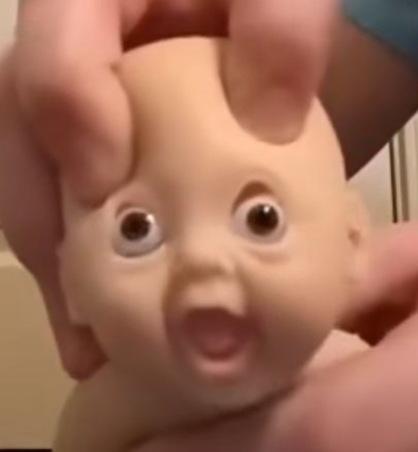 A hand squeezing a baby doll's head, its googly eyes and open mouth locked in shock