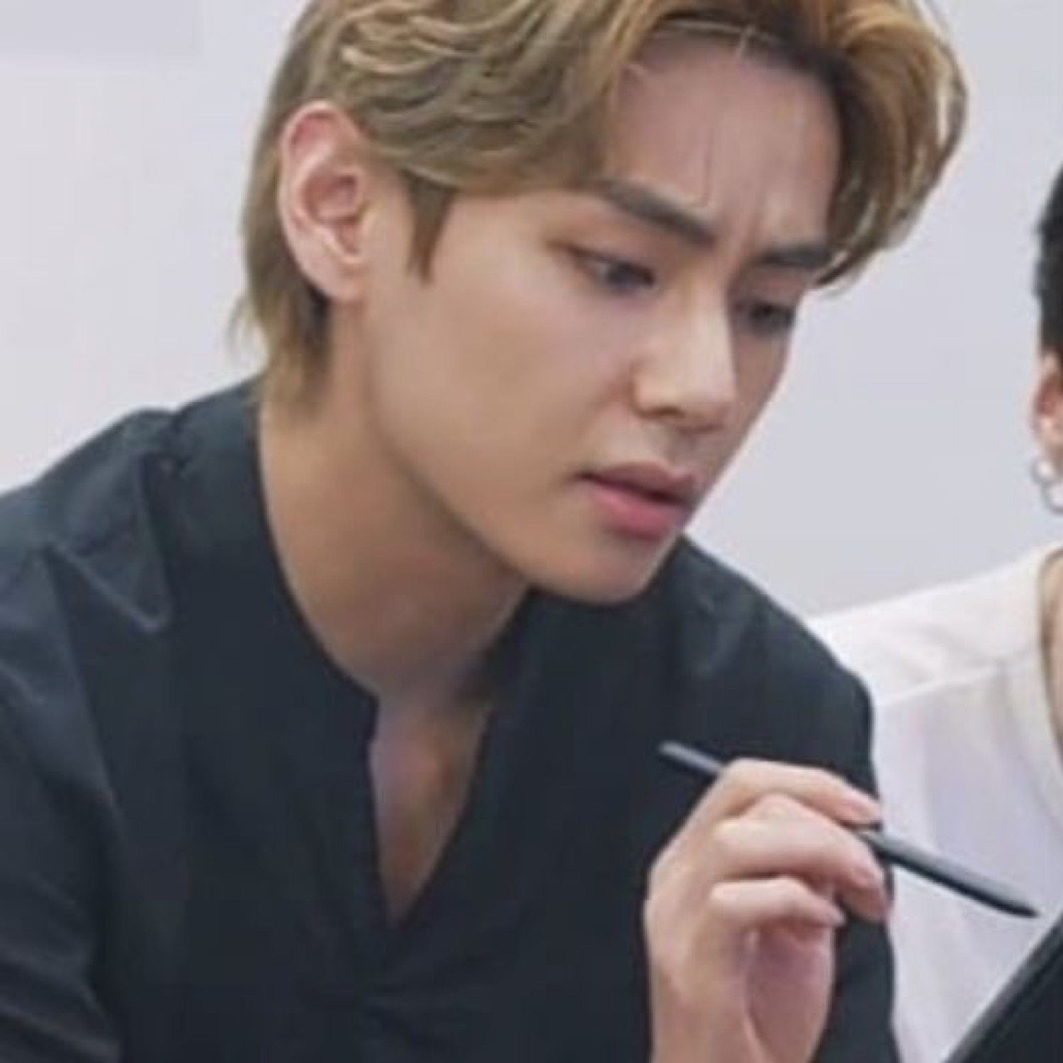 A blond man in black leaning in with a puzzled, focused frown, pen in hand