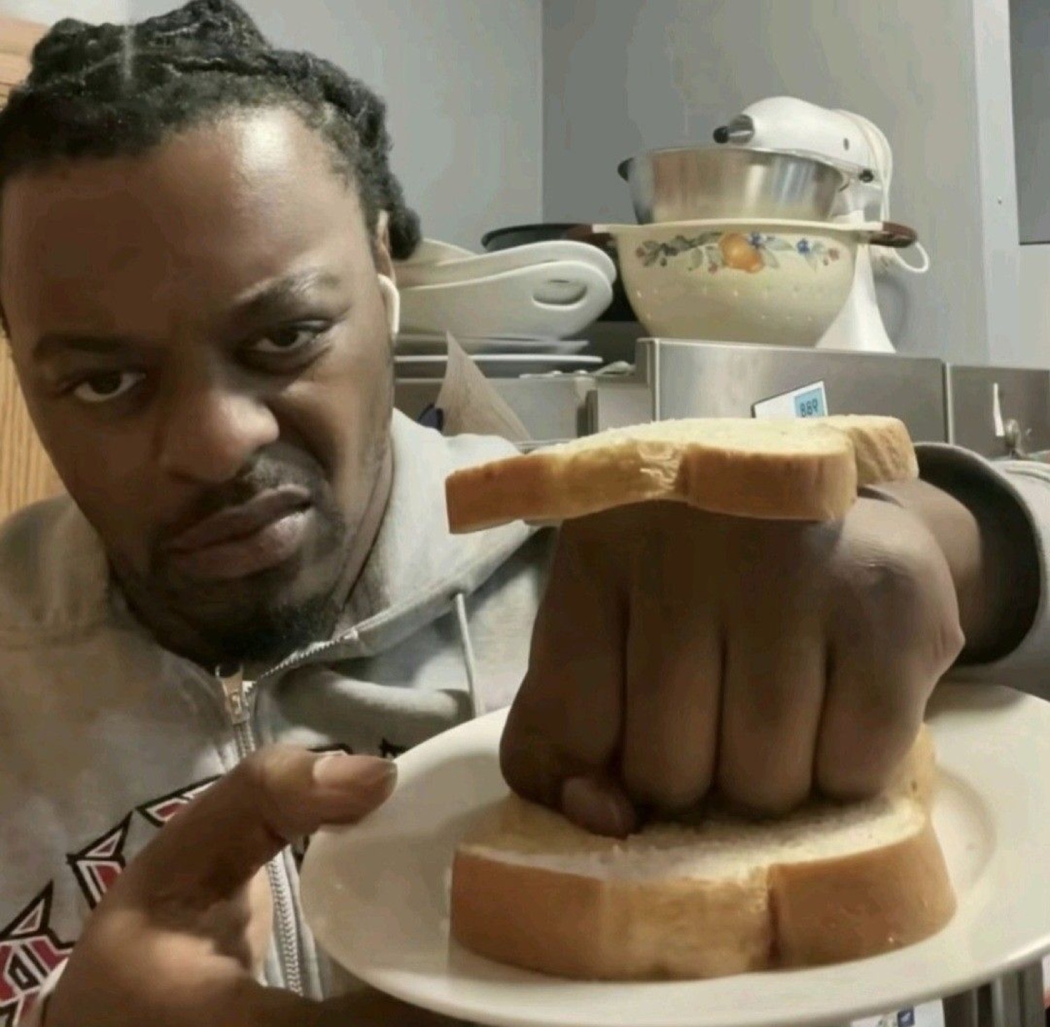 Man with a flat, unimpressed face pressing his fist into a sandwich of white bread on a plate