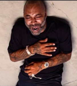 Bald tattooed man slumped against a wall clutching his chest, dying laughing