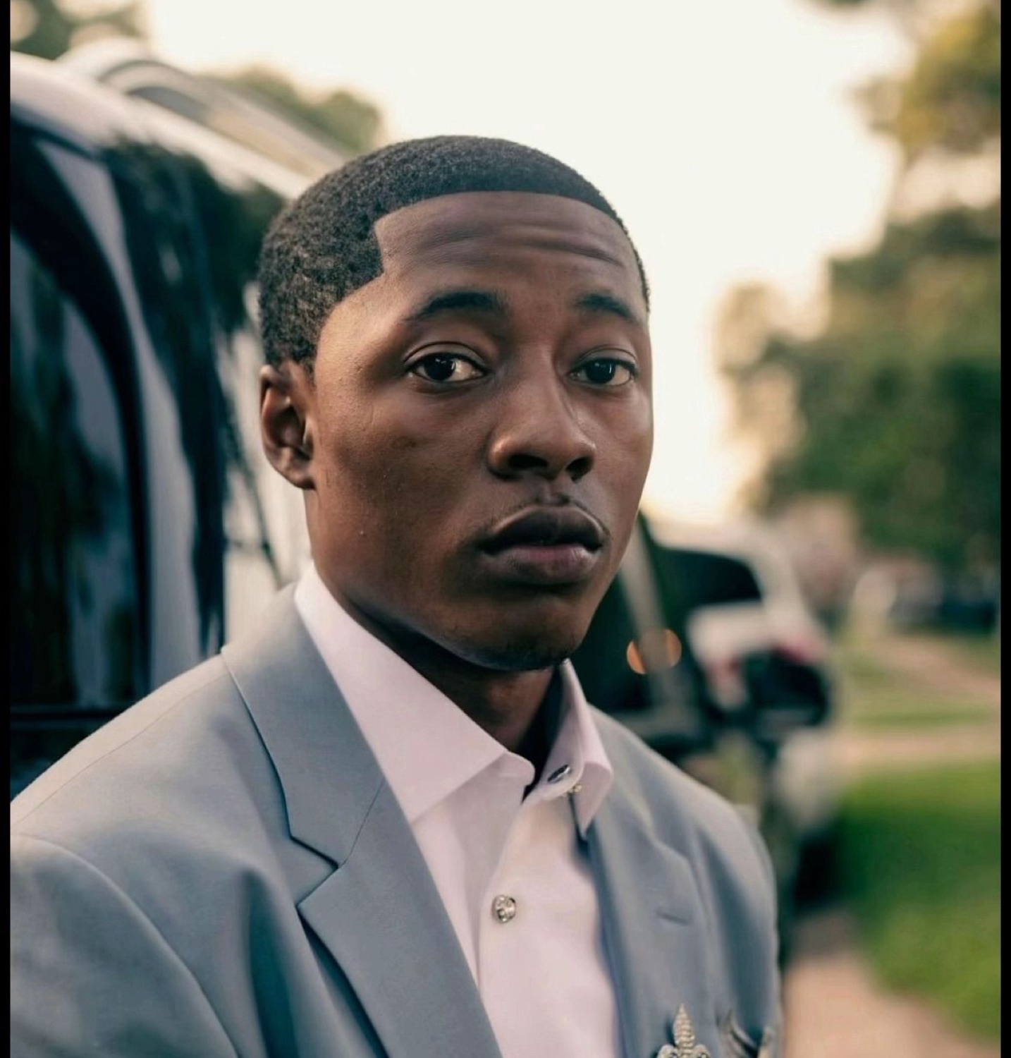 Young man in a grey suit leaning against a car giving a flat, unreadable stare