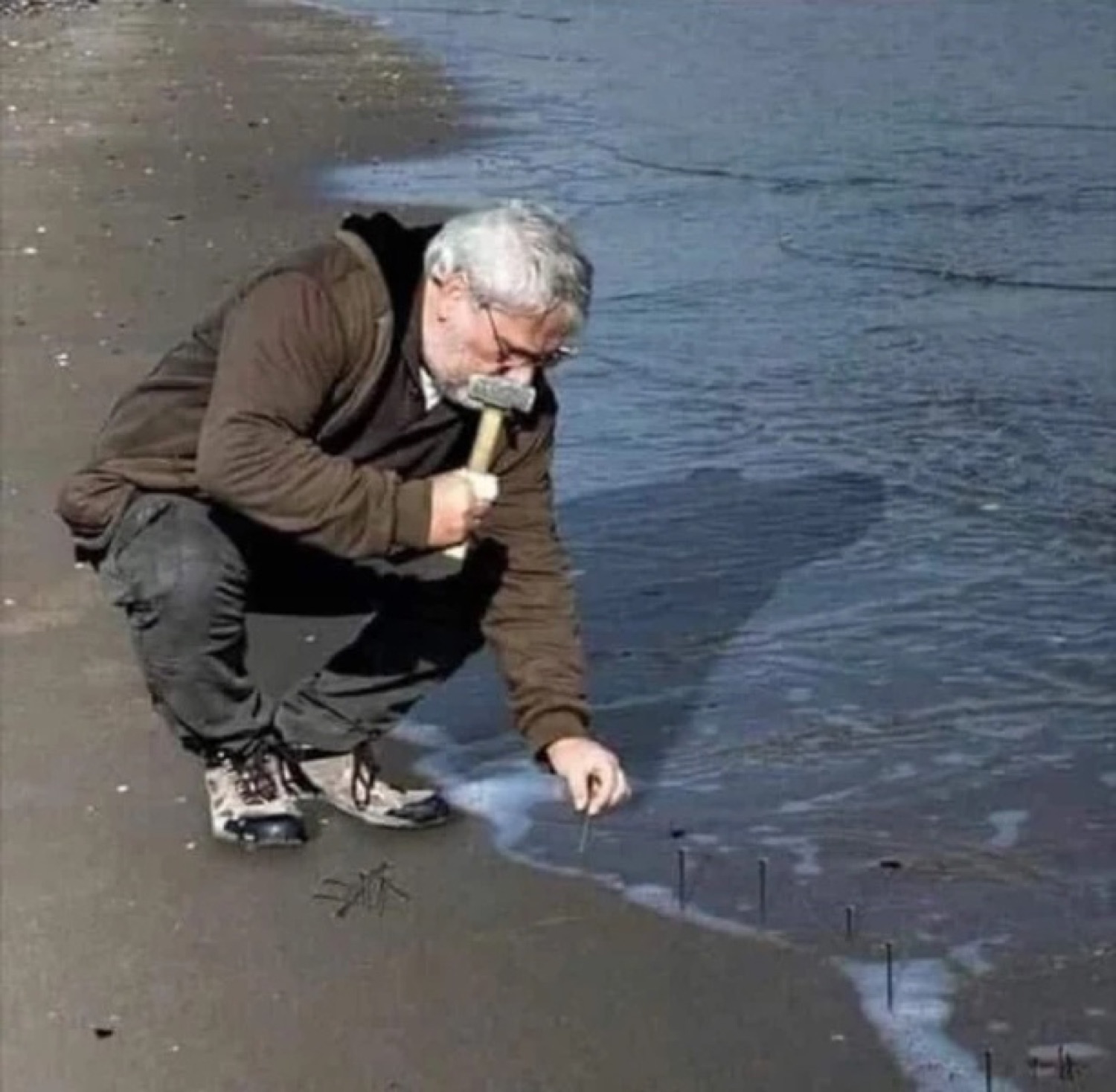 Man crouched at the shoreline hammering nails into the wet sand as the tide comes in