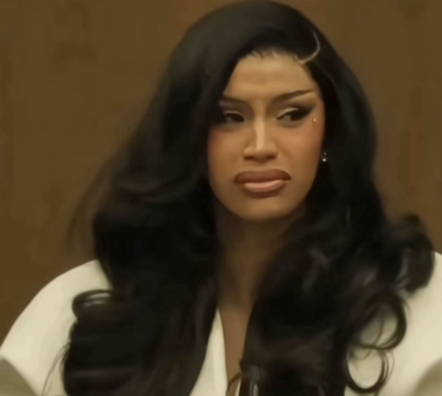 Woman with long black hair and a cool stare sitting at what looks like a courtroom table