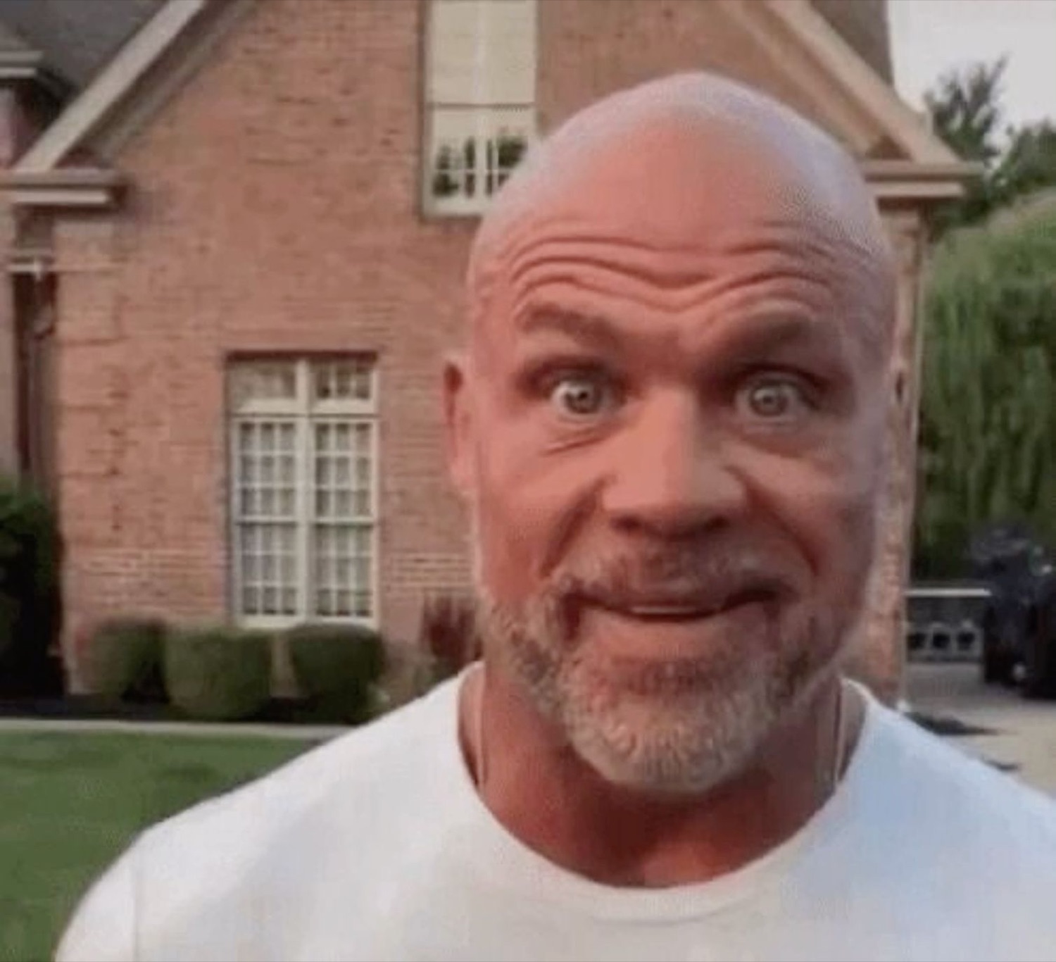 Bald man outside a brick house with wide, startled eyes and a stiff pursed smile