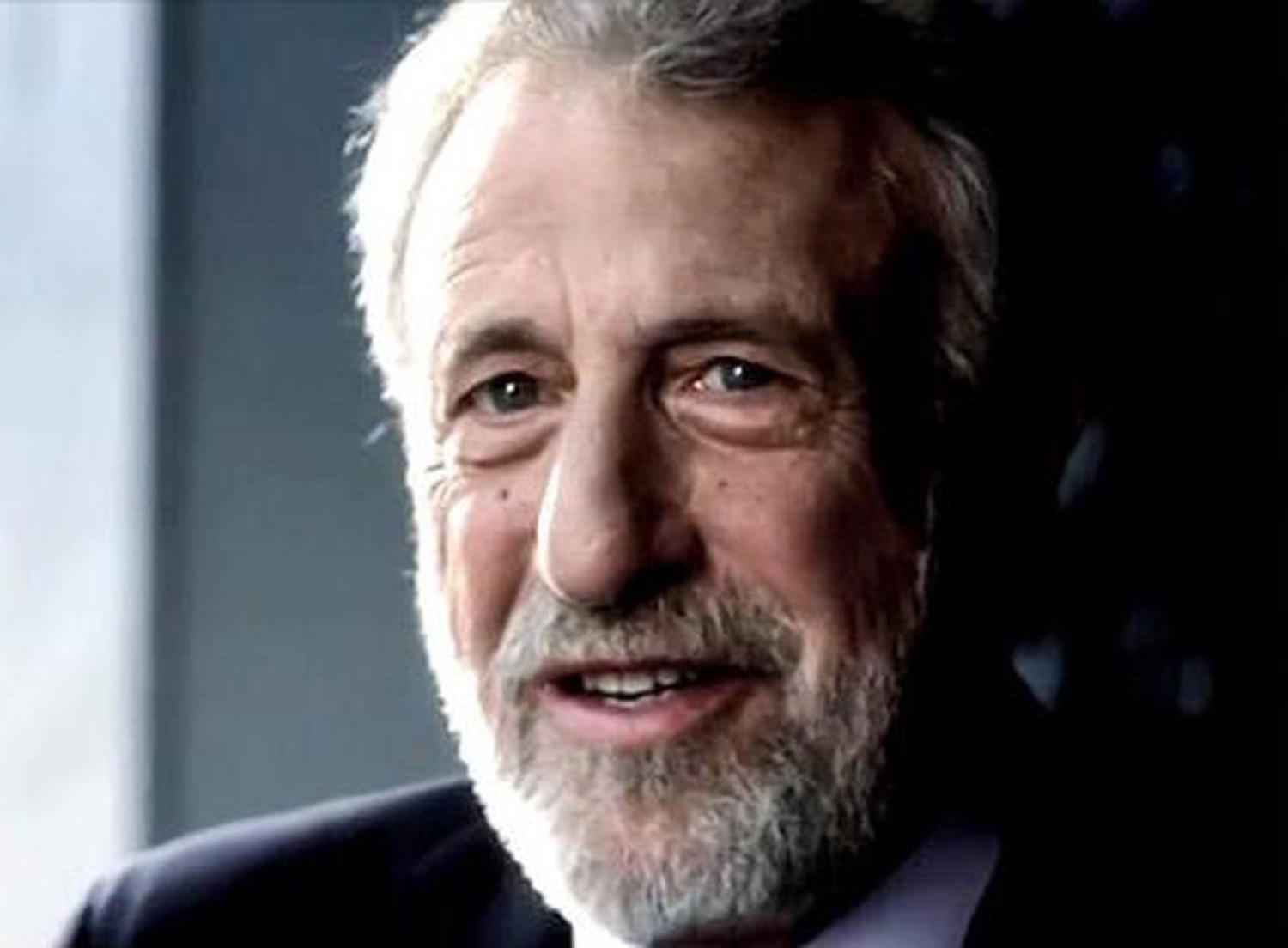 Silver-haired older man flashing a knowing smug smile in a dark suit
