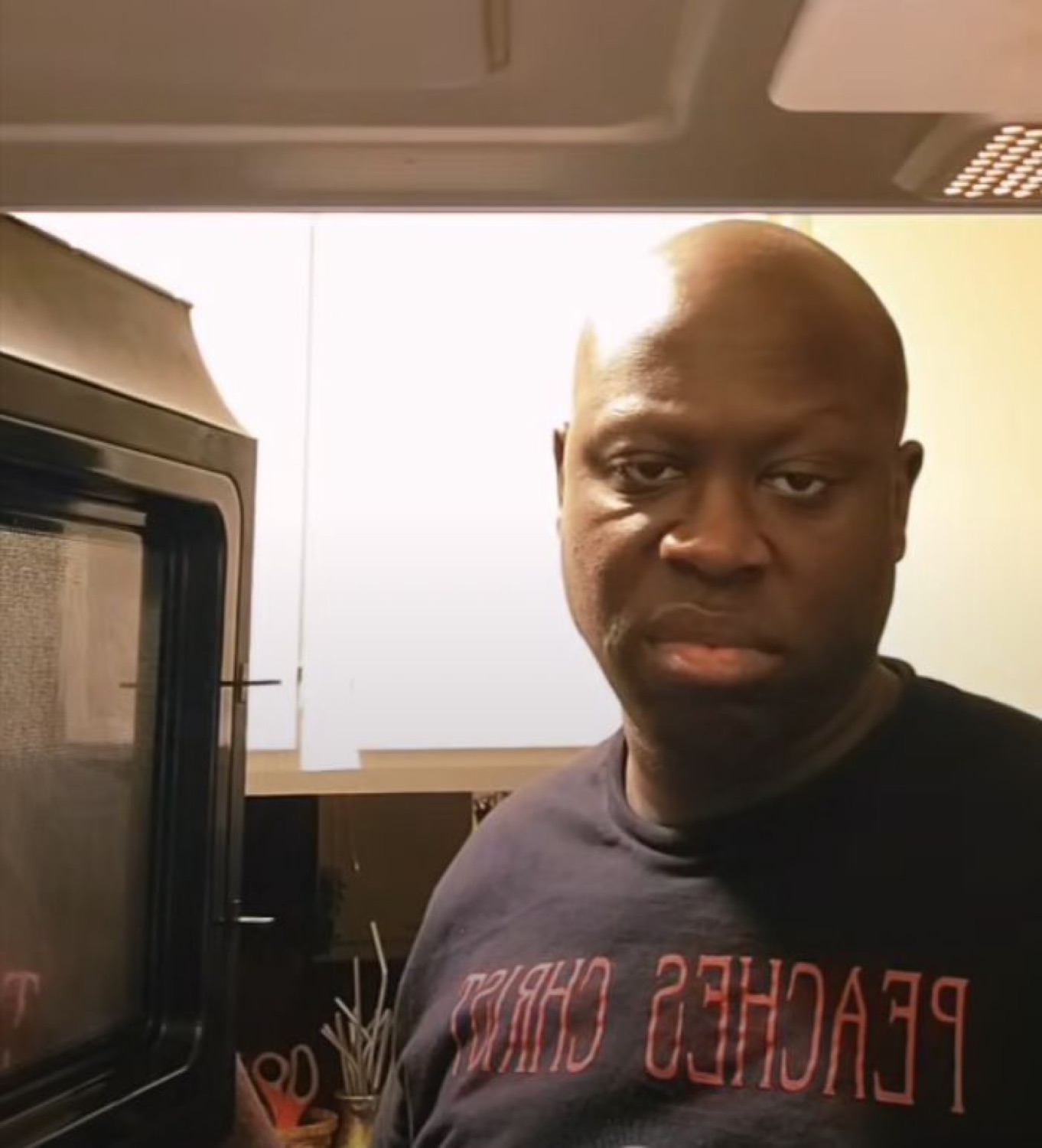 Bald man in a Peaches Christ shirt staring into the camera next to a microwave with zero emotion