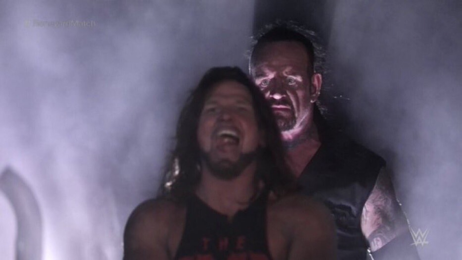 Wrestler laughing while a menacing figure looms behind him in the smoky dark