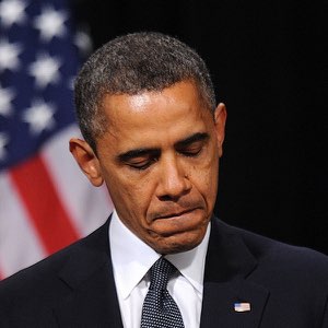Barack Obama looking downward with a heavy, disappointed frown in front of an American flag