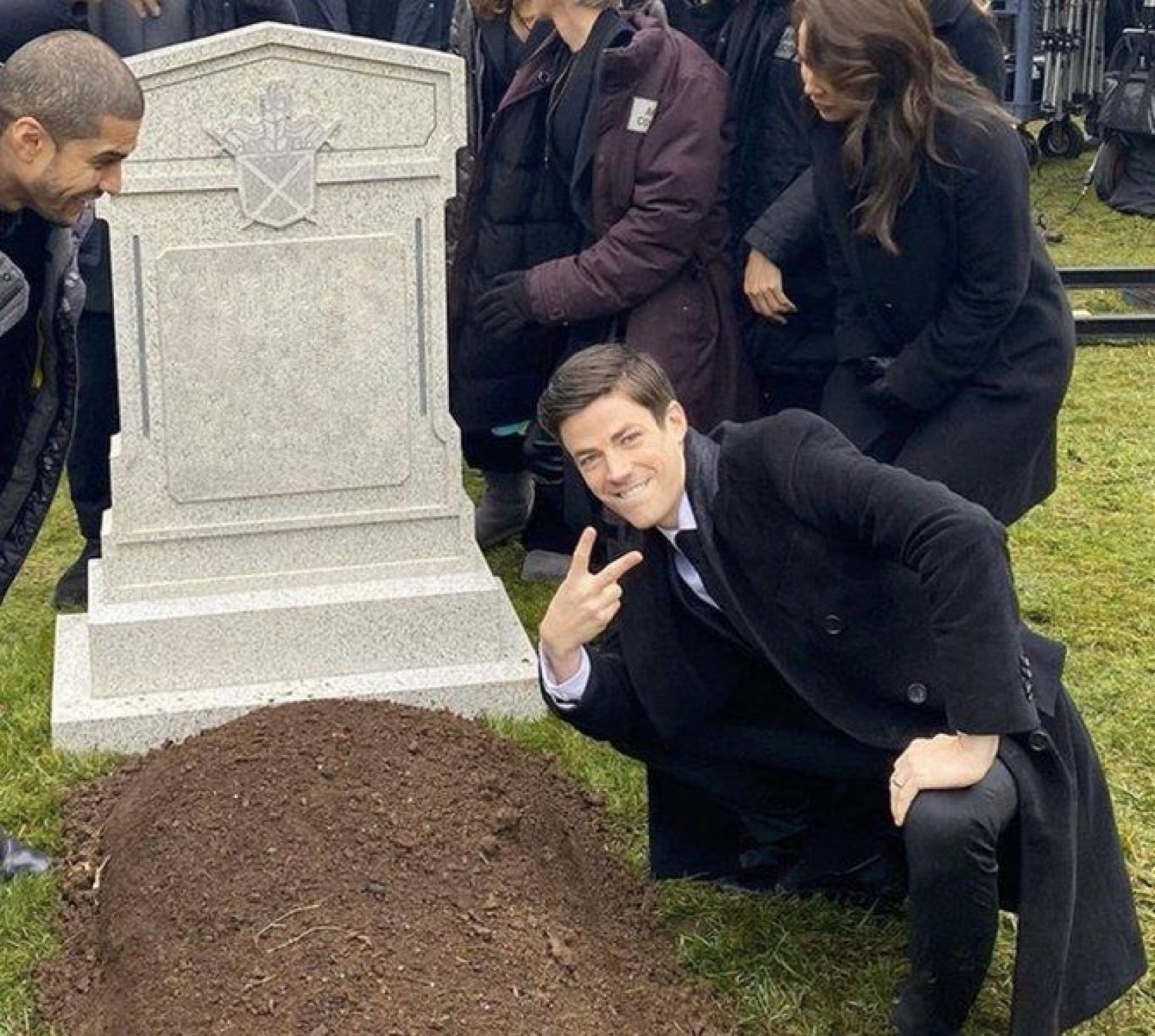 Man in a suit crouching by a fresh grave flashing a peace sign and grinning