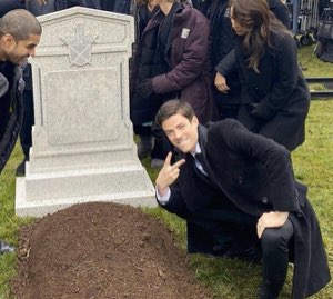 Man in a suit crouching by a fresh grave flashing a peace sign and grinning