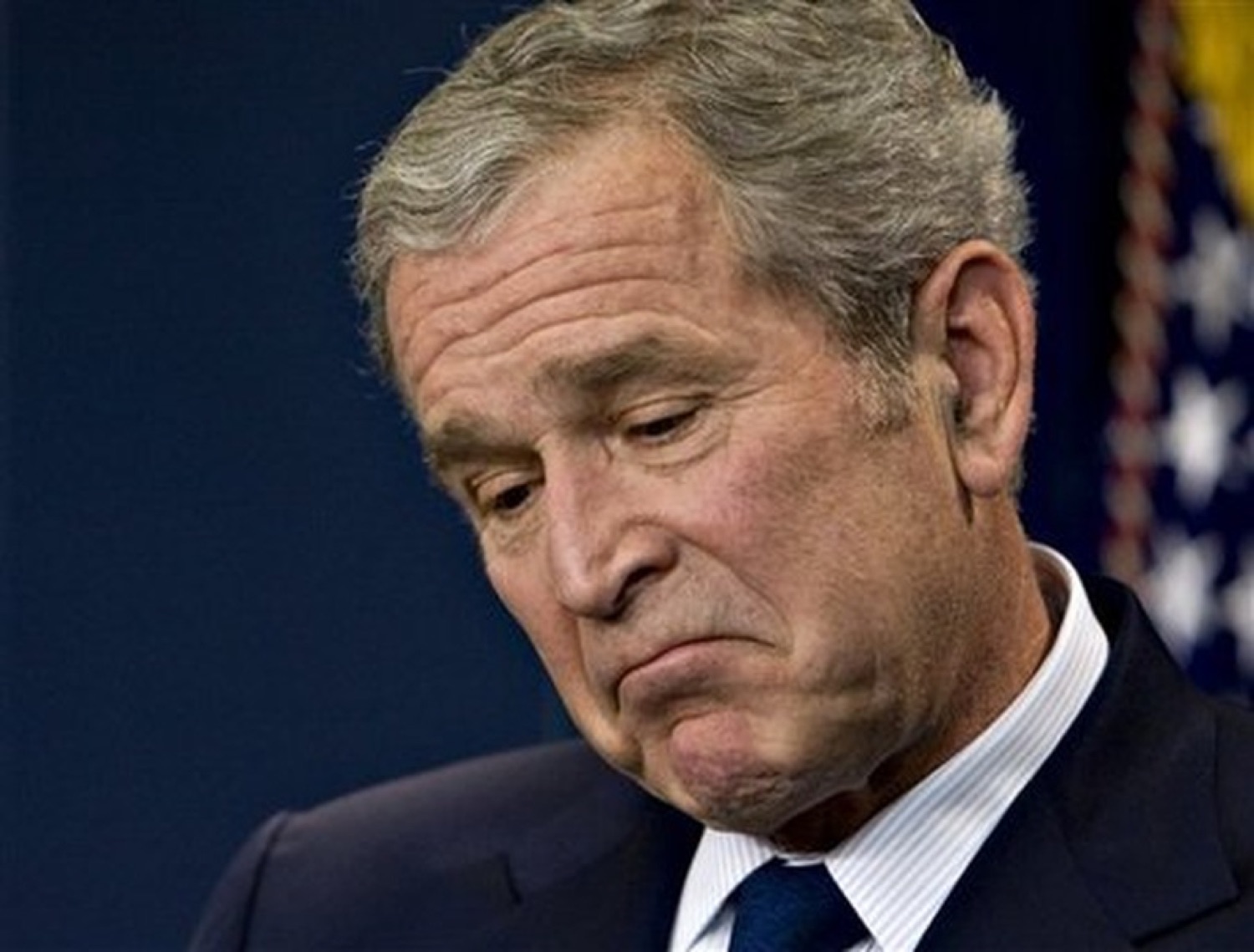 A man in a suit looking down with a heavy, disappointed frown in front of an American flag backdrop