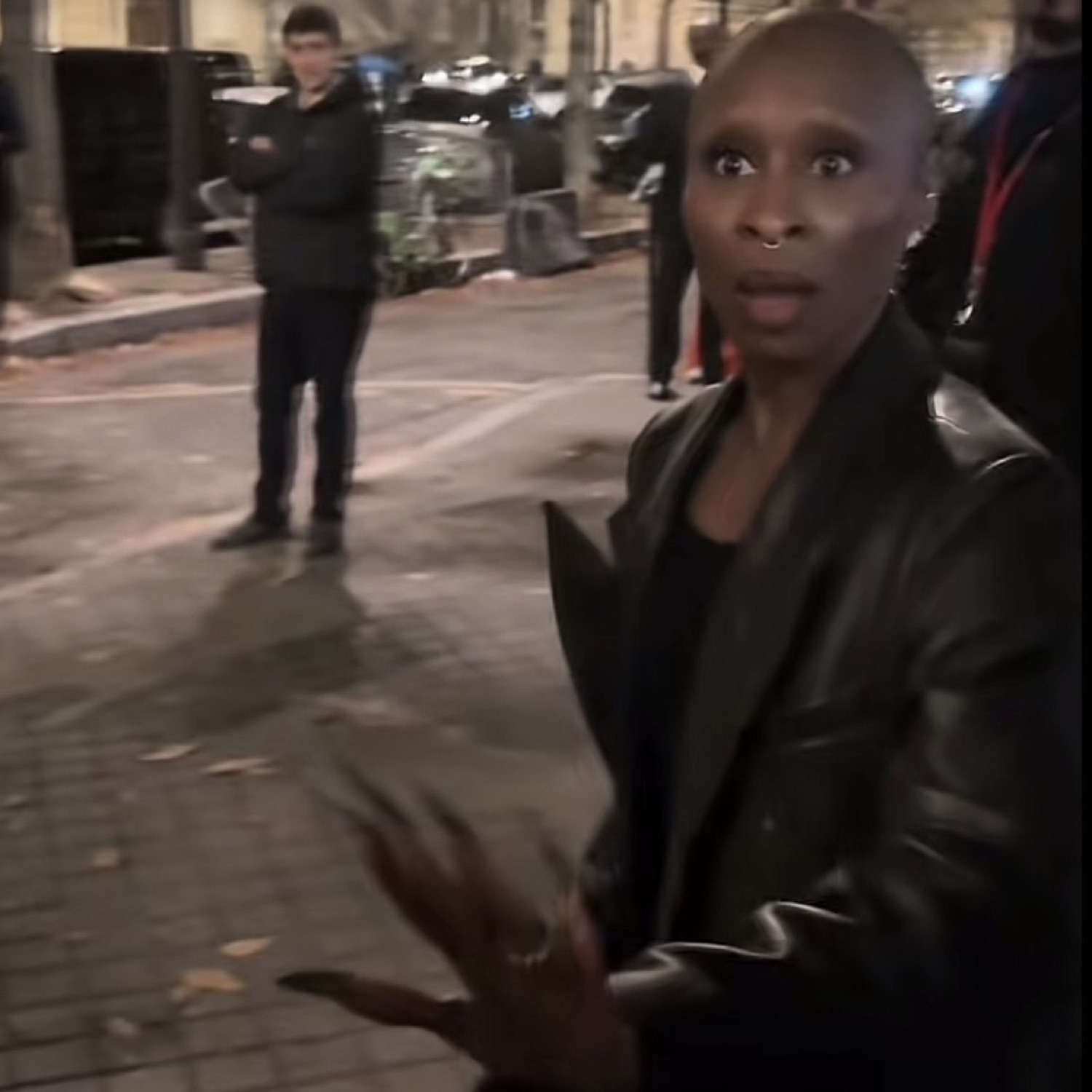 Bald woman in a leather jacket looking stunned with hands gesturing outward on the street