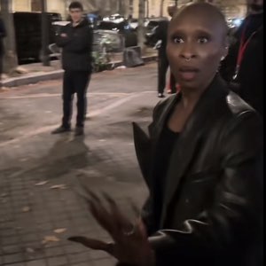Bald woman in a leather jacket looking stunned with hands gesturing outward on the street