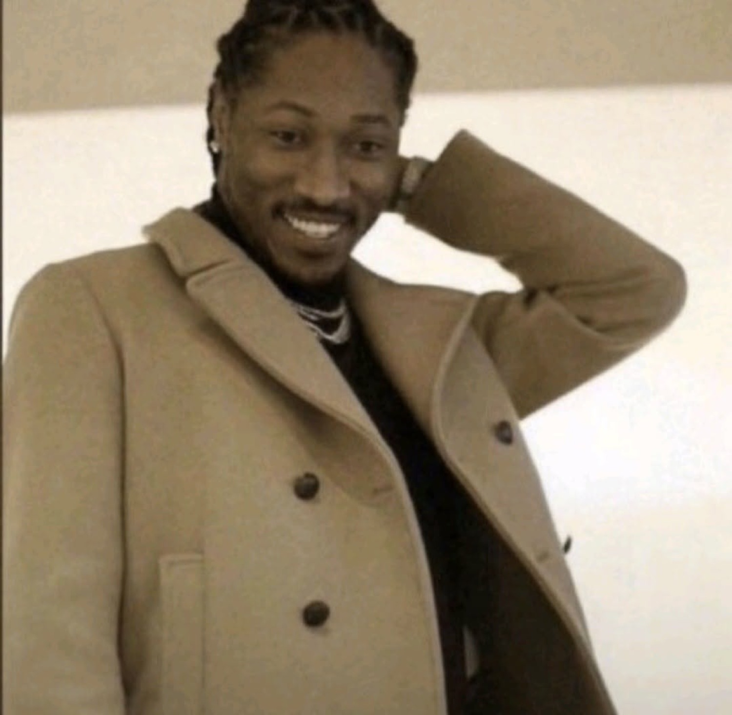 Man in a tan coat smiling smoothly with a hand behind his head, posing confidently