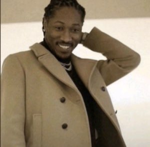 Man in a tan coat smiling smoothly with a hand behind his head, posing confidently