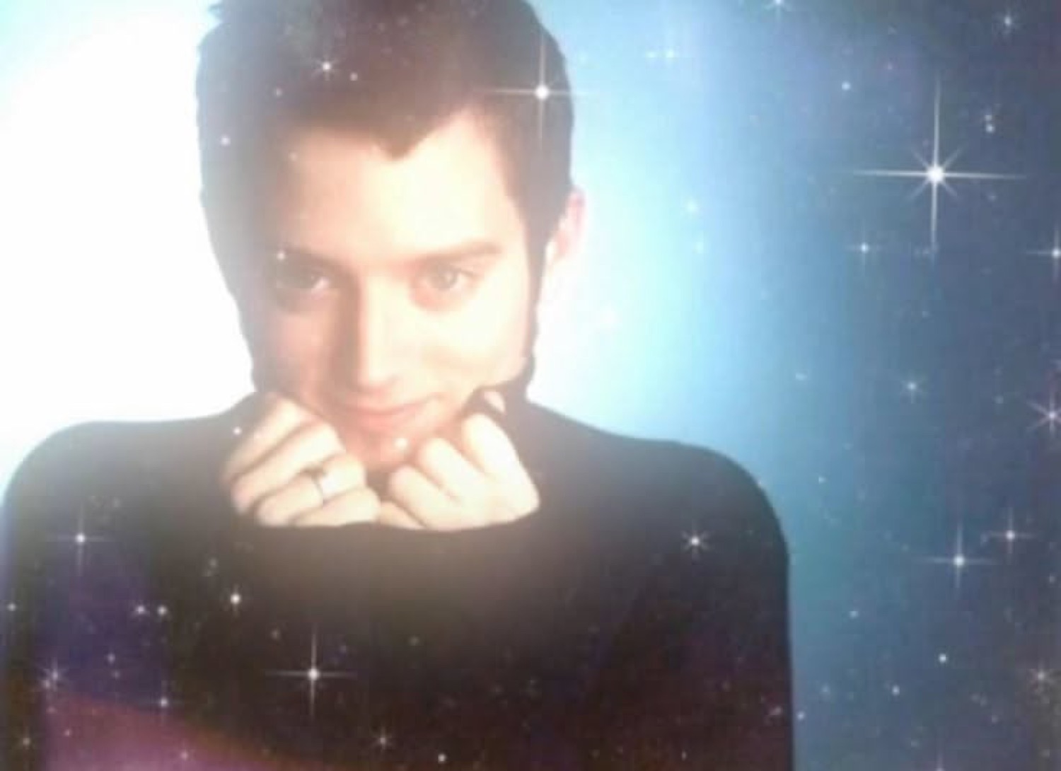 Man in a black turtleneck posing coyly with chin in hands against a sparkly star backdrop