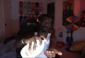 Man in headphones and a tank top reaching toward the camera with a deadpan look in his room