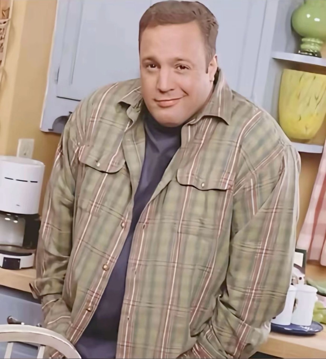 Heavyset man in a plaid shirt leaning on a kitchen counter with a satisfied smirk
