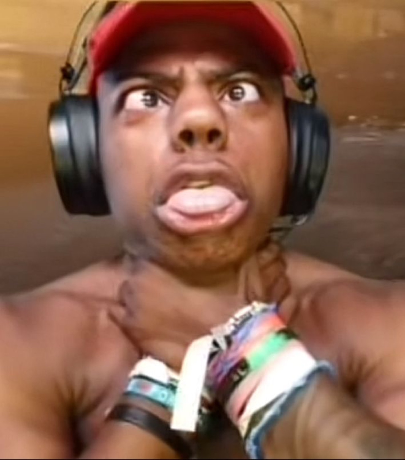 Man in a red cap and headphones with eyes rolled back and tongue out, gripping his neck