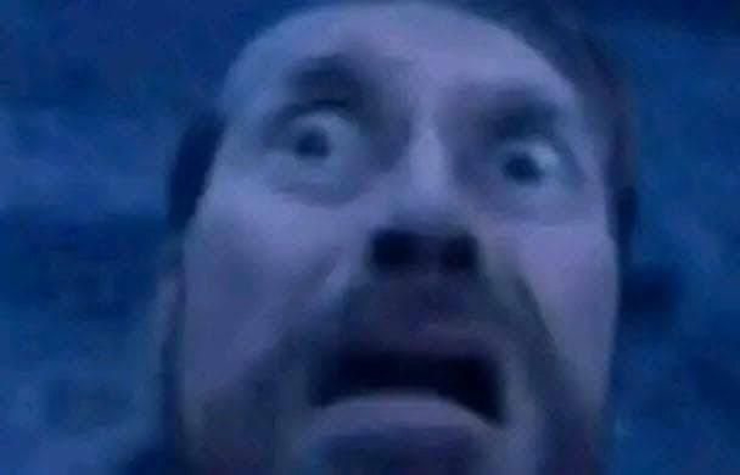 Blurry blue-lit close-up of a man with bulging eyes and open mouth, utterly terrified