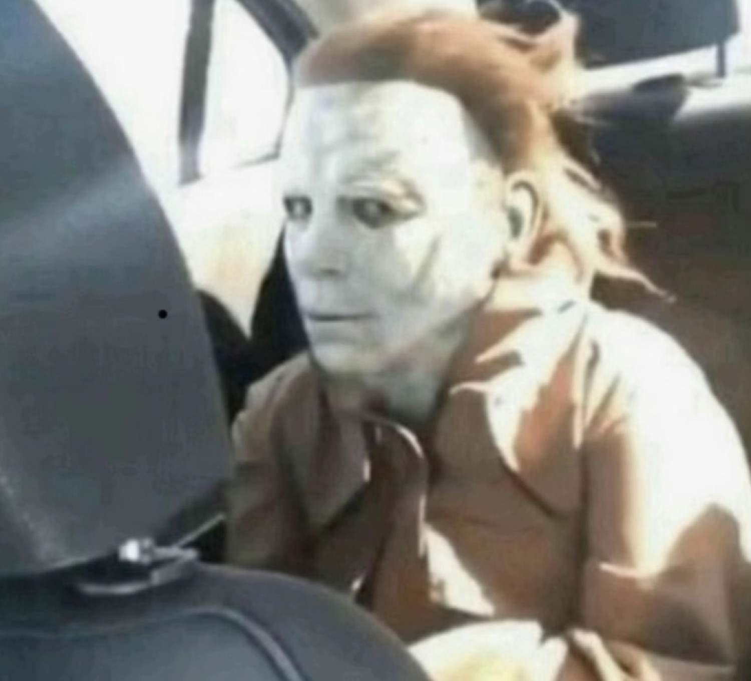 Person in a Michael Myers mask sitting in a car seat with a blank, deadpan stare