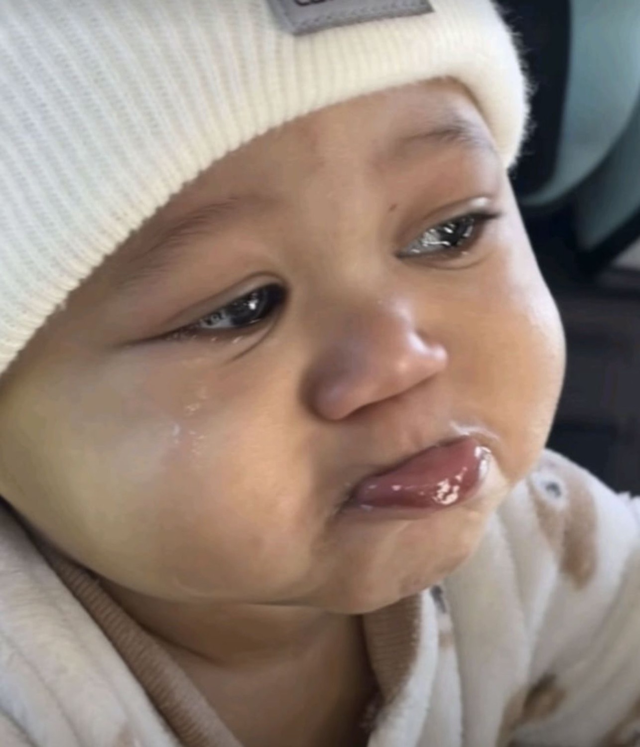 Baby in a cream beanie with tears streaming down, lip pushed out in a pout