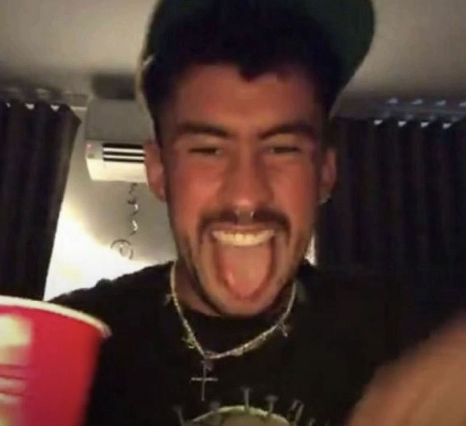 Guy in a backwards cap sticking his tongue out with a red cup at a party