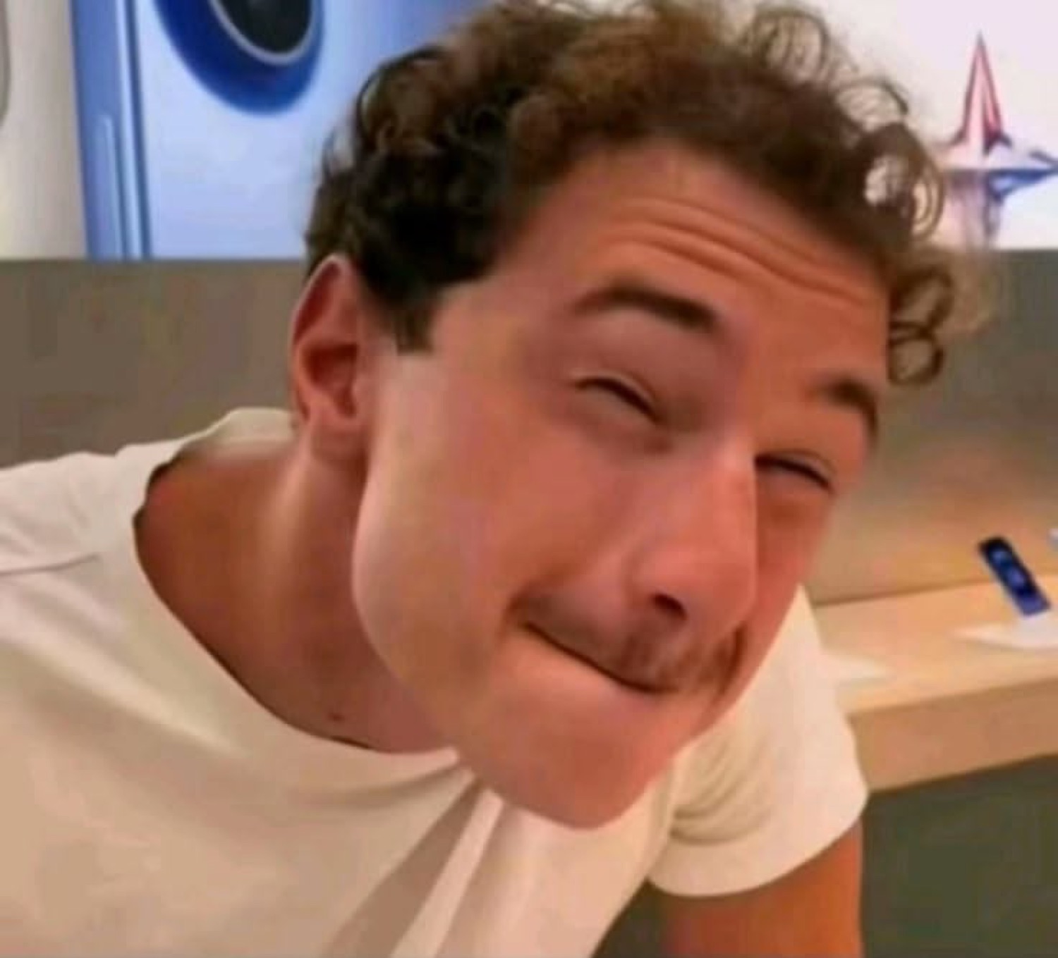 Curly-haired man leaning in with a squinty, awkward closed-mouth smile